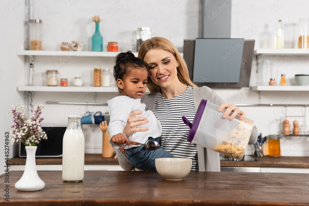 happy mother holding adopted african american kid with spoon and pouring corn flakes in bowl