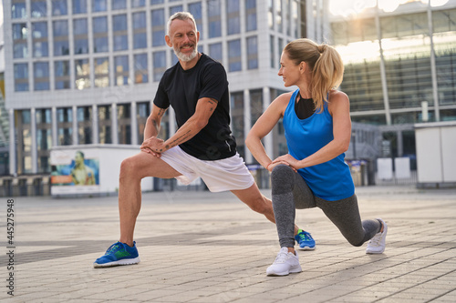 Active sporty middle aged couple, man and woman stretching legs, preparing for running together in the city on a summer day