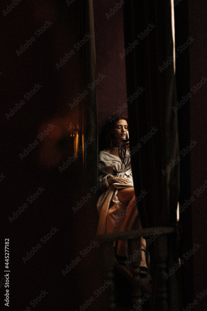 Cool lady sitting in dark room and drinking wine. Woman with red lips dressed in trench coat chilling in bar Cool lady sitting in dark room and drinking wine. Woman with red lips dressed in trench coat chilling in bar