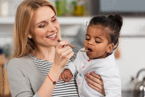 cheerful mother holding spoon and feeding adopted african american daughter