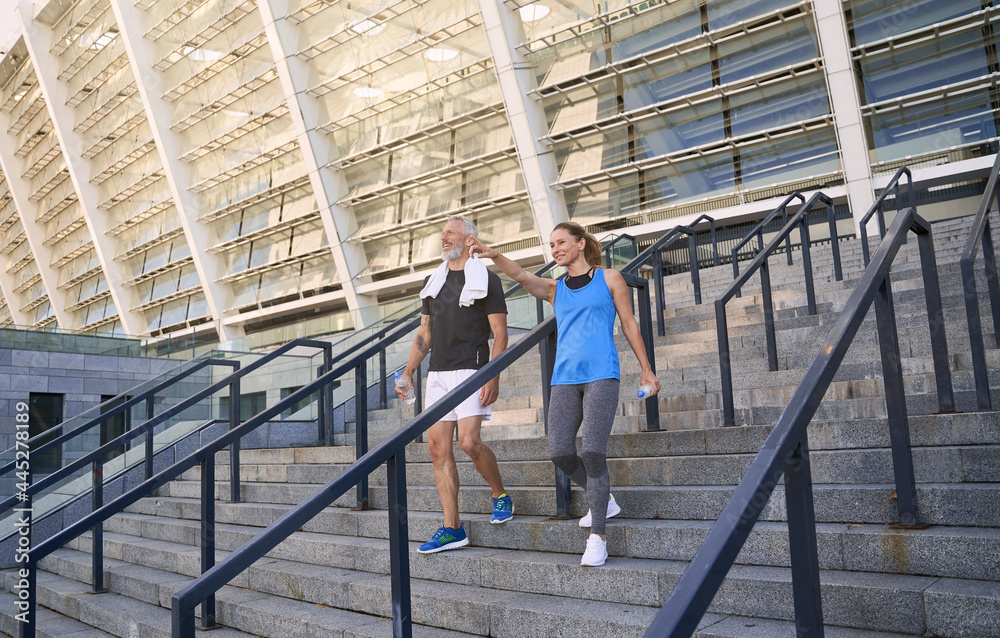 Active mature couple, man and woman in sportswear having conversation while walking down the stairs after training together outdoors