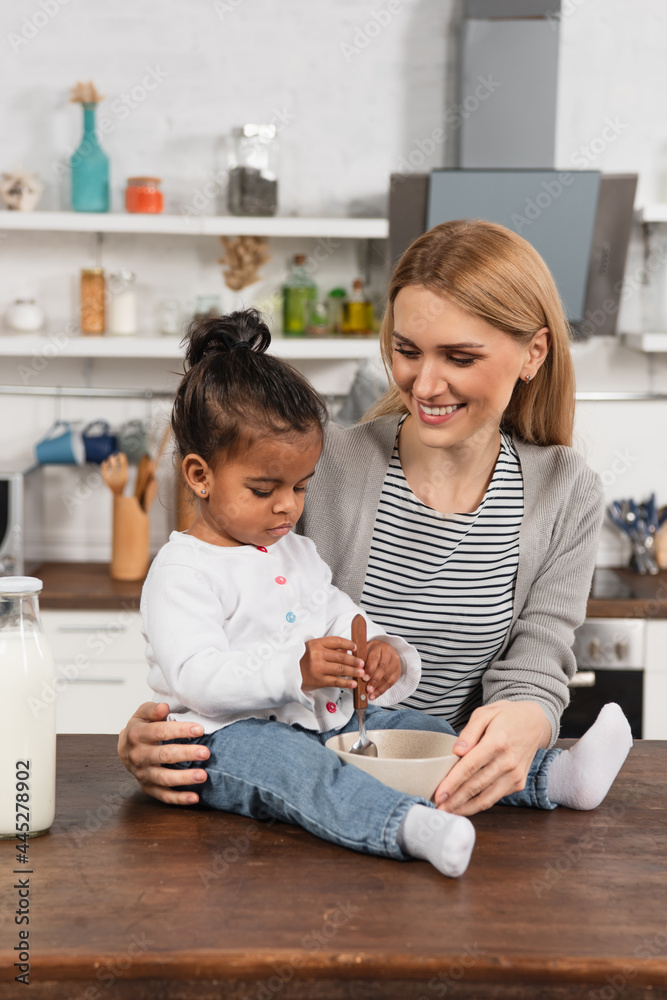 happy mother looking at adopted african american kid with spoon sitting on kitchen table near bowl happy mother looking at adopted african american kid with spoon sitting on kitchen table near bowl