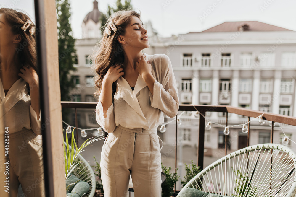 Curly woman enjoys sunny day on terrace. Pretty dark-haired lady in beige blouse and stylish pants poses on balcony - obrazy, fototapety, plakaty 