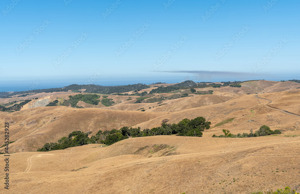 Cambria, CA, USA - June 9, 2021: View on blue ocean from Back country hills used for ranching under blue sky. Dry grass with patches of green trees. 