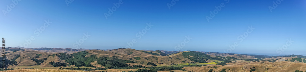 Cambria, CA, USA - June 8, 2021: Panorama landscape of ocean and Back country with dry ranch land and patches of green trees and agricultural fields under blue sky.