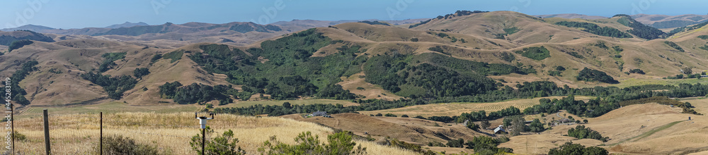 Cambria, CA, USA - June 8, 2021: Panorama landscape of hills and farm in Back country with dry ranch land and patches of green trees under blue sky.