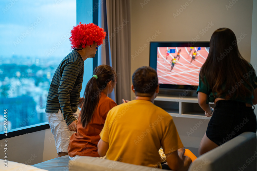 Fototapeta premium Group of Asian people friends sit on sofa watching and cheering sports games competition on TV together at home. Excited man and woman sport fans celebrate sport team victory in national sports match
