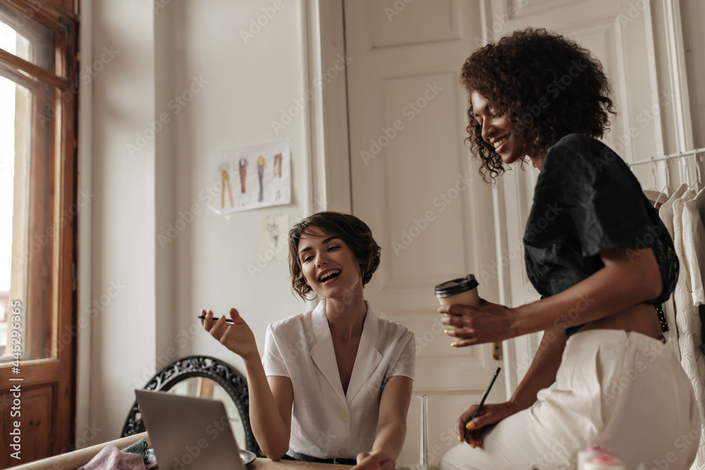 Fototapeta premium Pretty short-haired women in stylish blouses talk, smile and look at laptop screen in office. Curly dark-skinned woman in black top holds coffee cup.