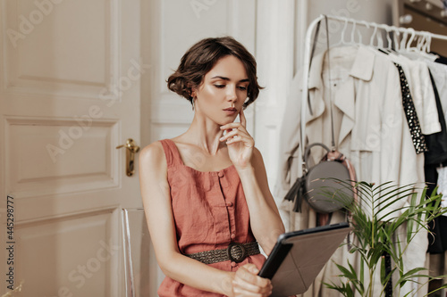 Thoughtful short-haired young woman in linen red dress sits on chair in cozy light room and holds computer table.