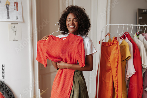 Excited young brunette curly dark-skinned woman in khaki shorts and white blouse smiles sincerely, holds hanger with red dress and poses in dressing room. photo