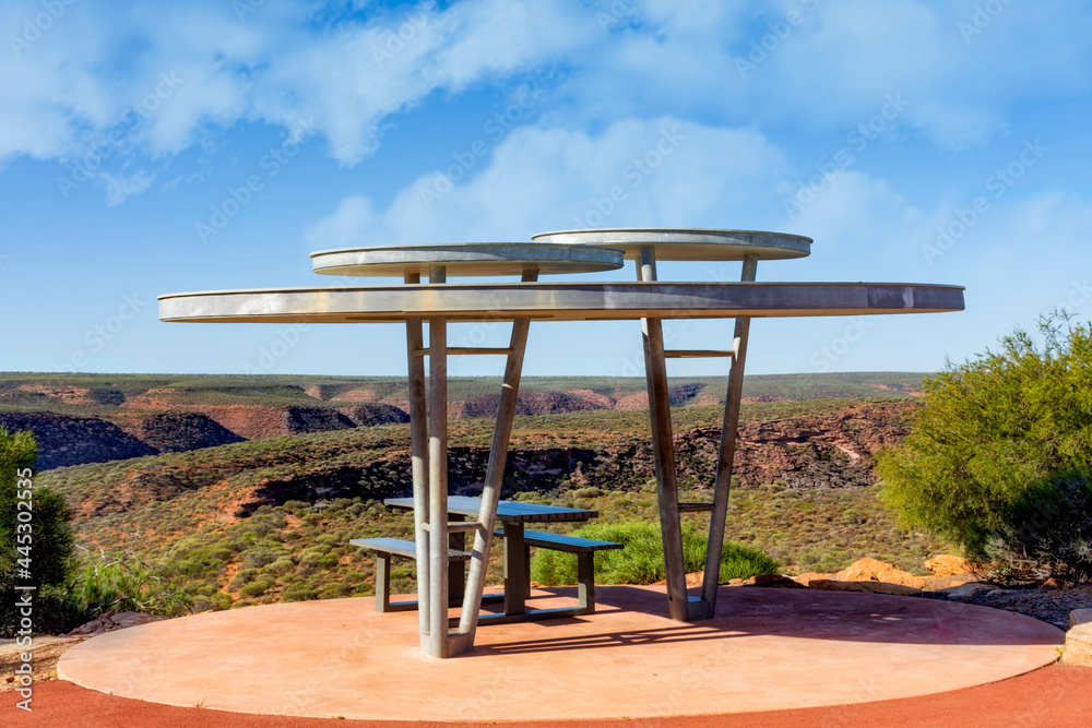 Shade shelter with benches set in the sandstone cliffs of the Kalbarri National Park surrounded by native flora under a blue sky with clouds in Western Australia. Shade shelter with benches set in the sandstone cliffs of the Kalbarri National Park surrounded by native flora under a blue sky with clouds in Western Australia.
