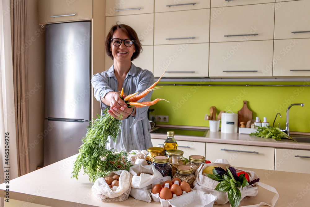 Adult woman stands the kitchen and holds out bunch of young fresh carrots. Concept of vegetarian food.