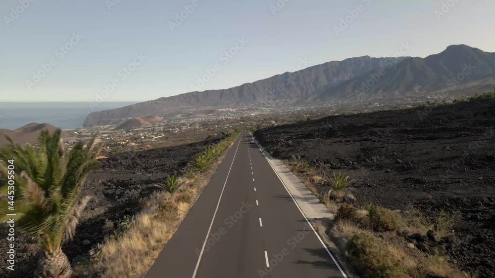 Car driving towards drone flying over a long, endless road located in a beautiful, fertile volcanic landscape Car driving towards drone flying over a long, endless road located in a beautiful, fertile volcanic landscape