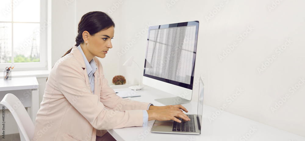 Accounting. Accountant Woman in the office of an audit company working with business documents. Serious woman in light pink suit working on desktop and laptop computers in white office. 