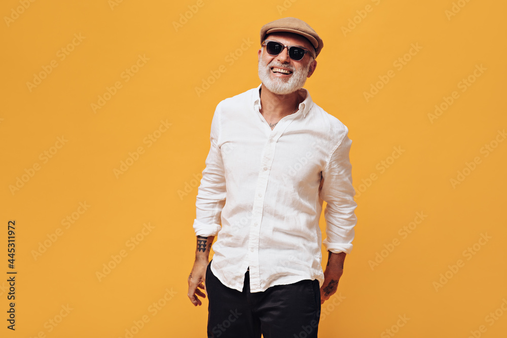 Charming man in cap, eyeglasses and white shirt smiling on orange background. Smiling adult in sunglasses in tattoos laughs into camera