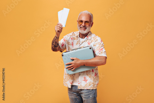 Charming man in plant print shirt poses with tickets and suitcase on orange background. Gray-haired guy with beard in summer clothes is having fun