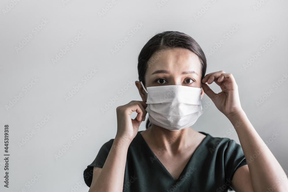 Asian woman wearing protection face mask against coronavirus. Studio shot masked woman looking at the camera and putting mask on her face on grey background. Coronavirus or Covid-19 concept. Asian woman wearing protection face mask against coronavirus. Studio shot masked woman looking at the camera and putting mask on her face on grey background. Coronavirus or Covid-19 concept.