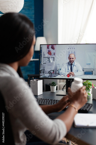 Fototapeta Naklejka Na Ścianę i Meble -  African american student holding pills box in hands while physician doctor explaining healthcare treatment against respiratory sickness. Patient discussing diagnosis during online videocall meeting