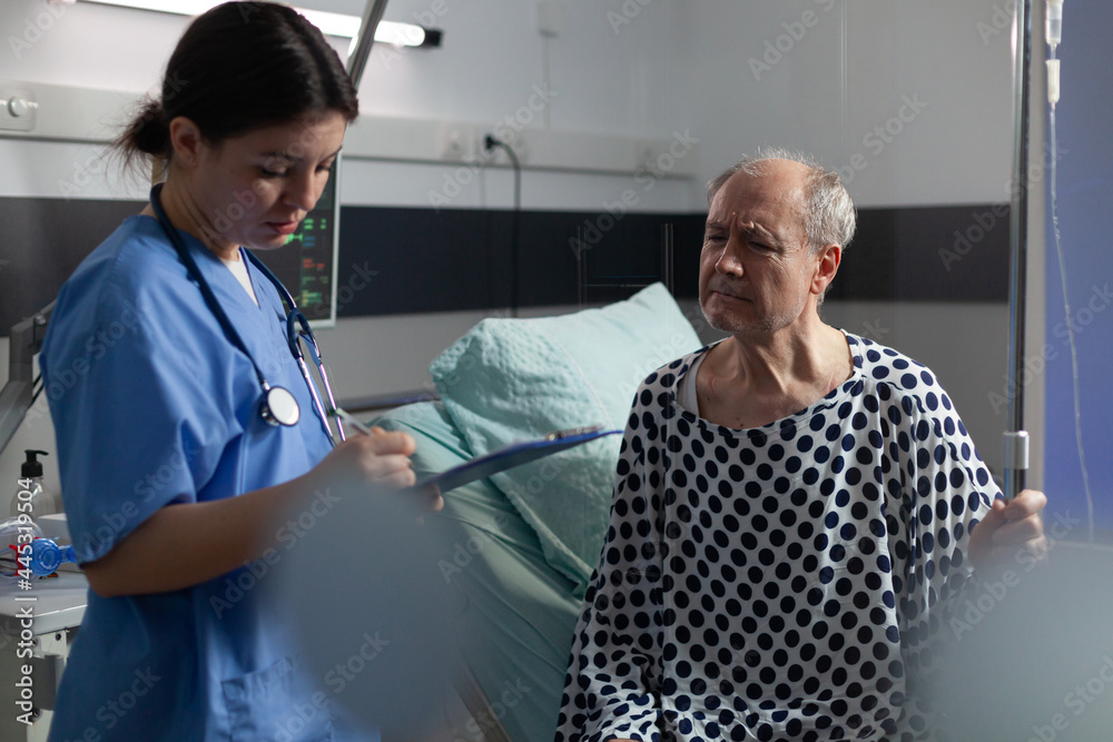Fototapeta premium Senior sick man in hospital room discussing with nurse about treatment during consultation. Medical staff with stethoscope writing notes on clipboard preparing patient for surgery.