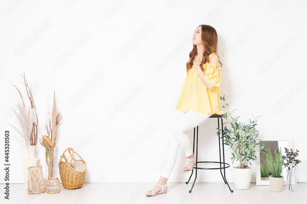 白い壁の前に置いてある椅子に座って、胸に手を当てる若い女性 白い壁の前に置いてある椅子に座って、胸に手を当てる若い女性