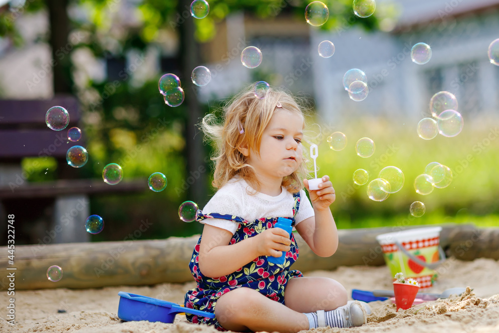 Beautiful little blonde toddler girl having fun with blowing soap bubble blower. Cute adorable baby child playing on playground on sunny summer day. Happy active funny healthy kid Beautiful little blonde toddler girl having fun with blowing soap bubble blower. Cute adorable baby child playing on playground on sunny summer day. Happy active funny healthy kid