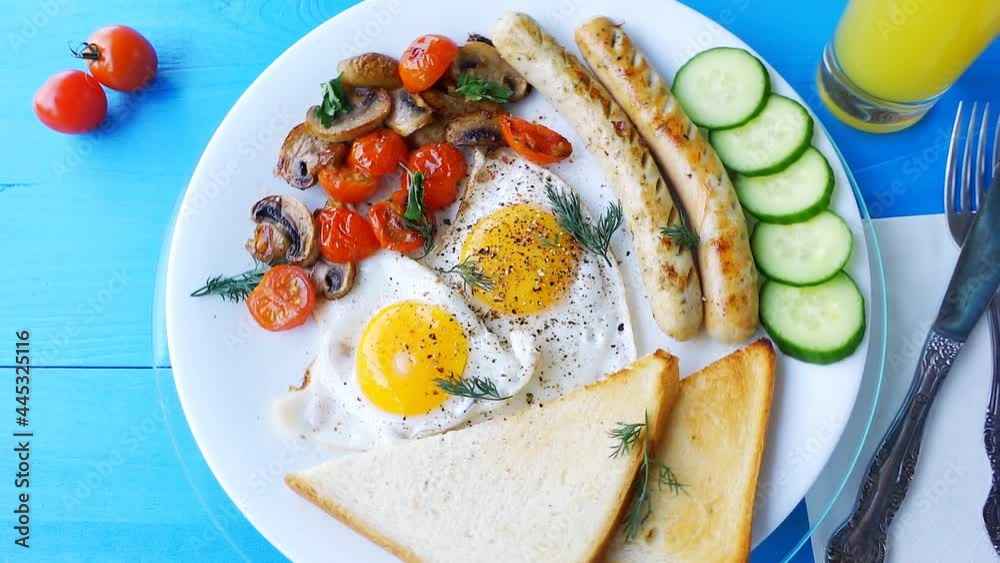Slow motion loop video of a breakfast of fried eggs, mushrooms, tomatoes, sausages and bread, rotating on a white plate on a wooden blue table. Slow motion loop video of a breakfast of fried eggs, mushrooms, tomatoes, sausages and bread, rotating on a white plate on a wooden blue table.