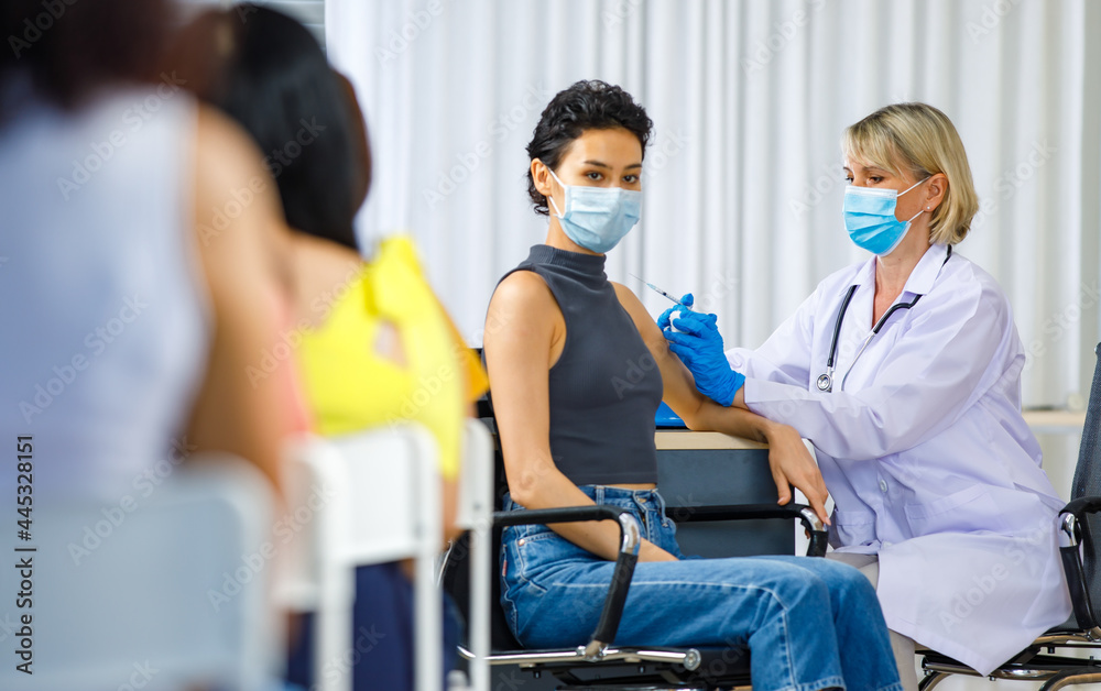 Obraz premium Cute female wears face mask sit look at camera while Caucasian woman doctor in white lab coat blue gloves and stethoscope injecting shot vaccine on shoulder when other patients wait in queue line