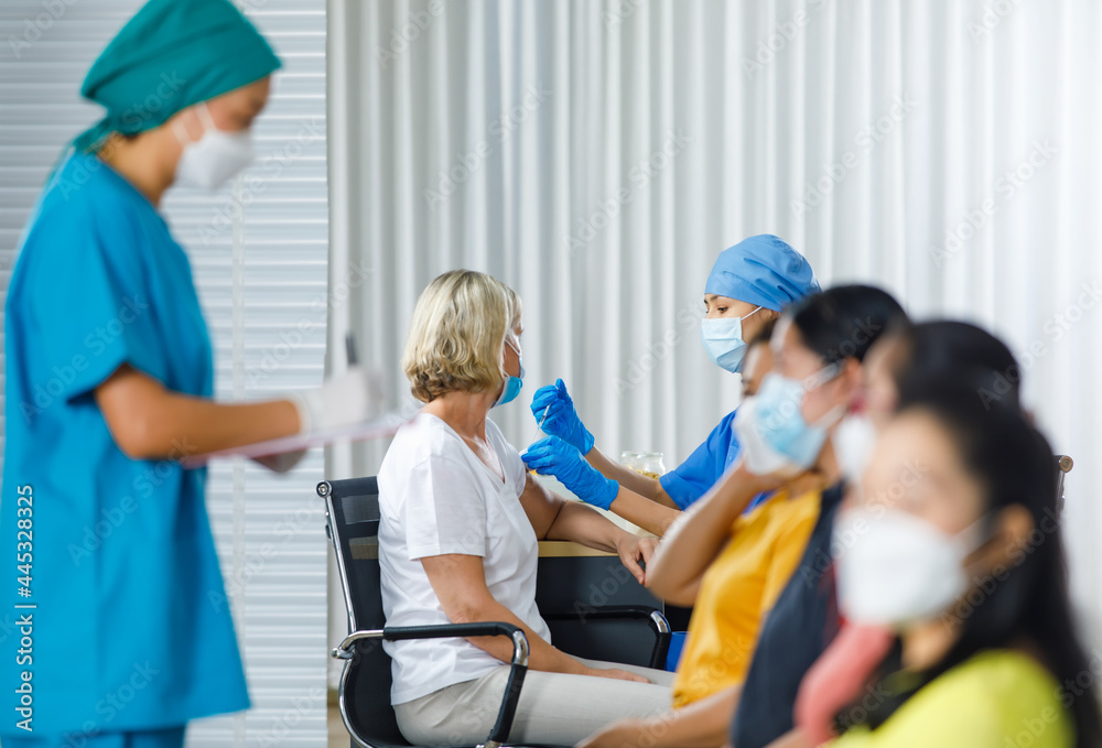 Doctor wears face mask injecting vaccine to Caucasian woman while nurse getting personal information from female patients sitting waiting in queue to covid 19 vaccinating in blurred foreground Doctor wears face mask injecting vaccine to Caucasian woman while nurse getting personal information from female patients sitting waiting in queue to covid 19 vaccinating in blurred foreground