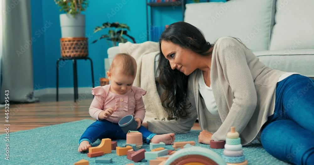 Funny, cute girl is playing with blocks with mother on carpet, she takes plastic container to her mouth, holds it, woman asks daughter to take it out, child lets go of toy and laughs