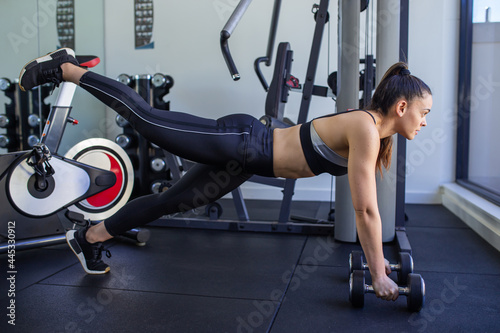 fitness woman wearing black outfit is training inside a small gym with black flooring, upside down lifting one leg.