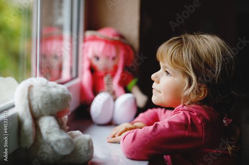Cute toddler girl sitting by window and looking out on rainy day. Dreaming child with doll and soft toy feeling happy. Self isolation concept during corona virus pandemic time. Lonely kid.