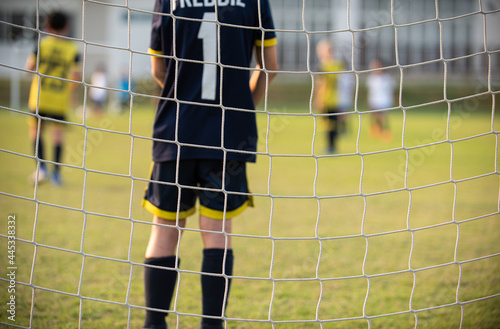 Close up of football goal net, seeing a young soccer goalie goalkeeper during the match. Youth Soccer game on a sunny summer school tournament Day. Junior football match going on in a background.
