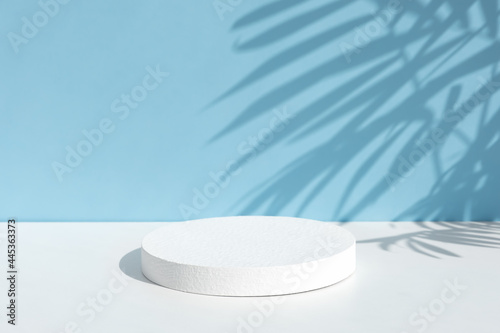Minimal abstract background for the presentation of a cosmetic product. A cylindrical white scene on a white table. Premium podium with a shadow of tropical palm leaves on a blue wall.