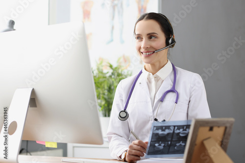 Doctor in headphones with microphone holding x ray in front of computer screen photo