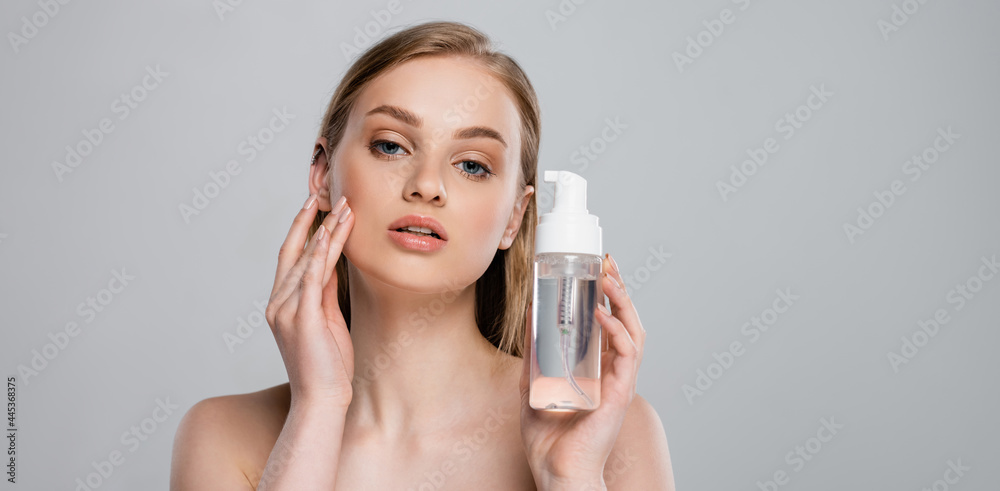 pretty young woman with bare shoulders holding bottle with micellar water isolated on grey, banner pretty young woman with bare shoulders holding bottle with micellar water isolated on grey, banner