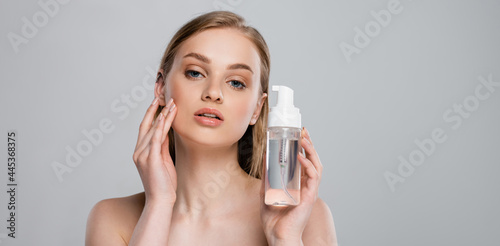 pretty young woman with bare shoulders holding bottle with micellar water isolated on grey, banner