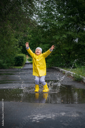 A little girl in a yellow raincoat and yellow boots is jumping in a puddle in a city park. Image with selective focus.