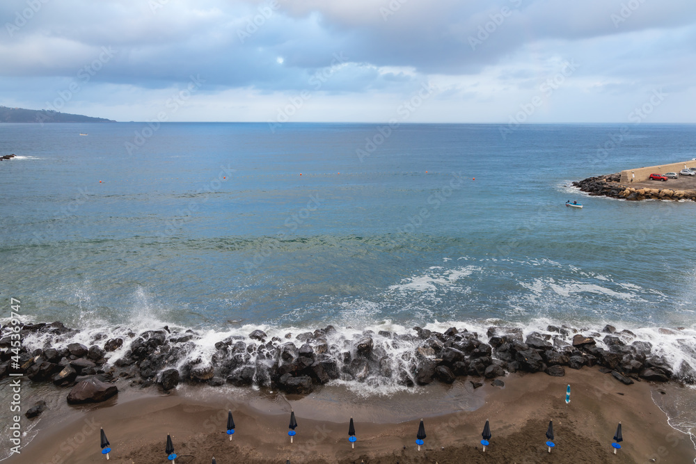 Big sea waves crash against the rocks next to beach full of umbrellas