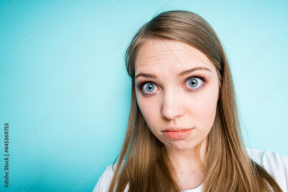 Beautiful cute girl in surprise looks at the camera raising her eyebrows and standing on a blue background Beautiful cute girl in surprise looks at the camera raising her eyebrows and standing on a blue background