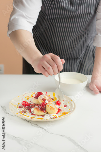 The chef in an apron serves a plate of cheesecakes. Cheesecakes are decorated with bright sauces and berries.