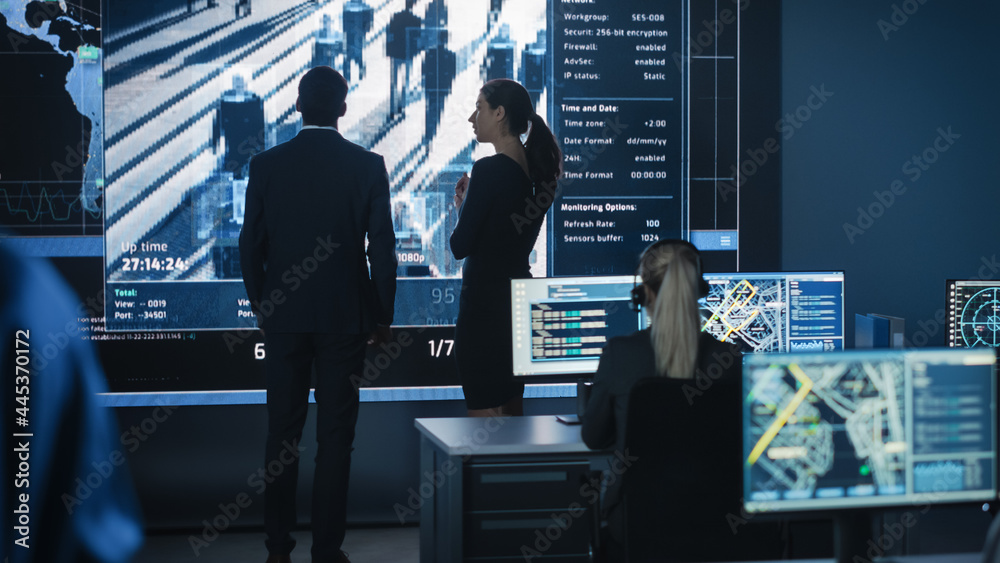 Male and Female Government Officers Discuss Working Matters next to a Computer and Big Digital Screen with Satellite Surveillance Footage with Face Recognition Software. Police Investigators at Work.