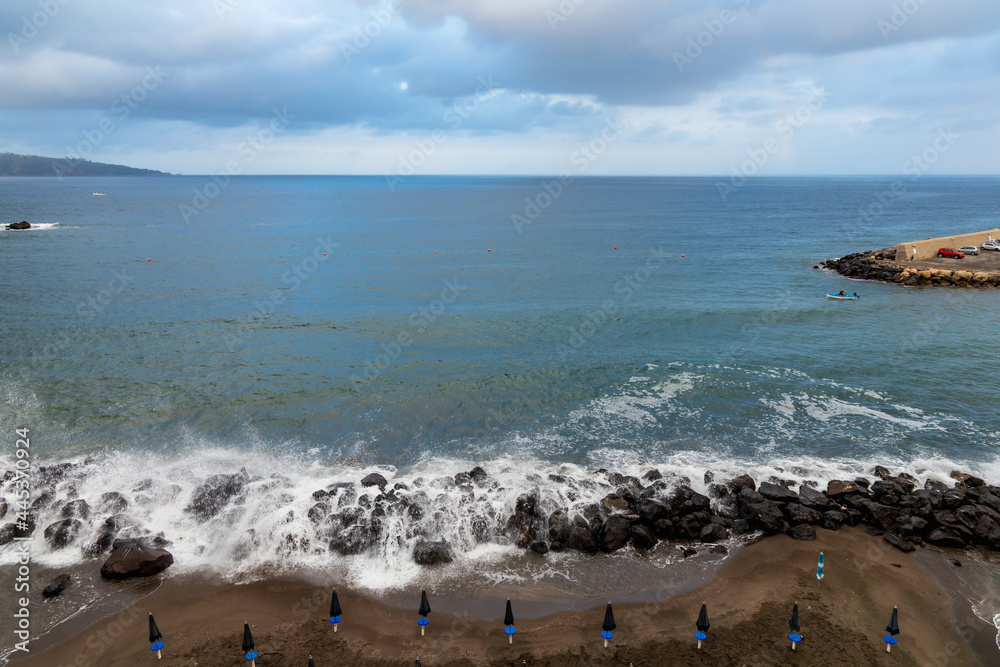 Big sea waves crash against the rocks next to beach full of umbrellas