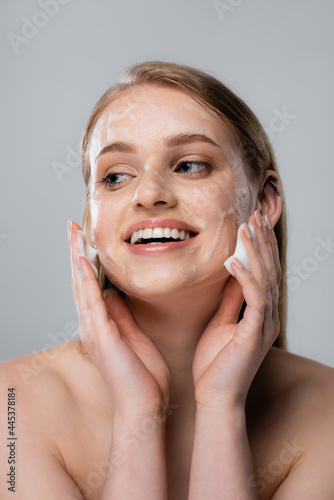 joyful young woman with blue eyes and cleansing foam on face isolated on grey