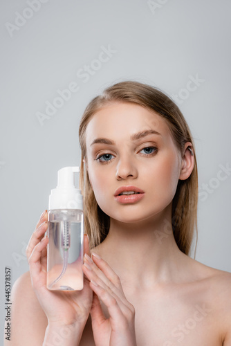 young woman with bare shoulders holding bottle with micellar water isolated on grey