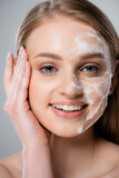 happy young woman with blue eyes and cleansing foam on face isolated on grey