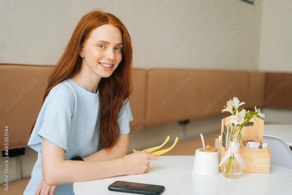 Medium shot portrait of smiling attractive young woman holding in hands yellow stylish eyeglasses sitting at table in cafe, looking at camera. Pretty redhead Caucasian lady posing in cozy coffee shop. Medium shot portrait of smiling attractive young woman holding in hands yellow stylish eyeglasses sitting at table in cafe, looking at camera. Pretty redhead Caucasian lady posing in cozy coffee shop.