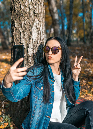 Young beautiful woman in sunglasses looks at the phone and takes a selfie, in the autumn park. Woman is resting in the autumn forest. Autumn time
