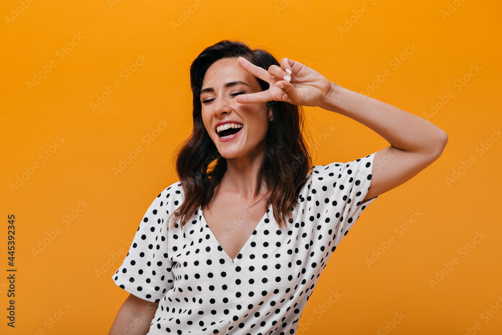 Naughty girl laughs and shows peace sign on orange background. Charming brunette in white polka-dot clothes is smiling and having fun Naughty girl laughs and shows peace sign on orange background. Charming brunette in white polka-dot clothes is smiling and having fun