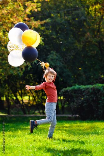 A child of primary school age with balloons in an autumn sunny park. A red-haired boy with a bunch of multicolored gold, black, and silver balls.
