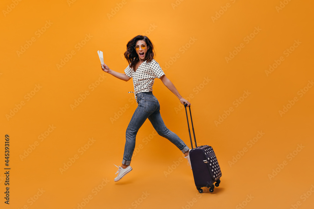 Beautiful girl in jeans is jumping on orange background. Full-length portrait of woman with tickets and suitcase Beautiful girl in jeans is jumping on orange background. Full-length portrait of woman with tickets and suitcase